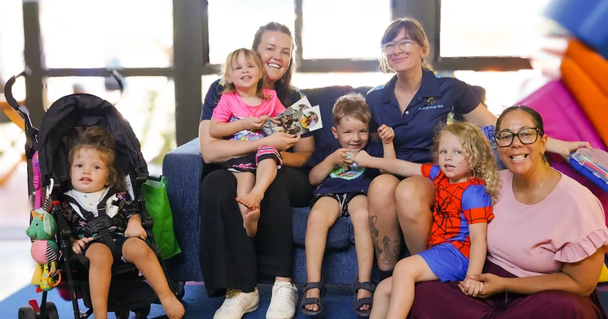 Three adults sitting on a couch, with three children in their laps and a toddler in a pram on the side 