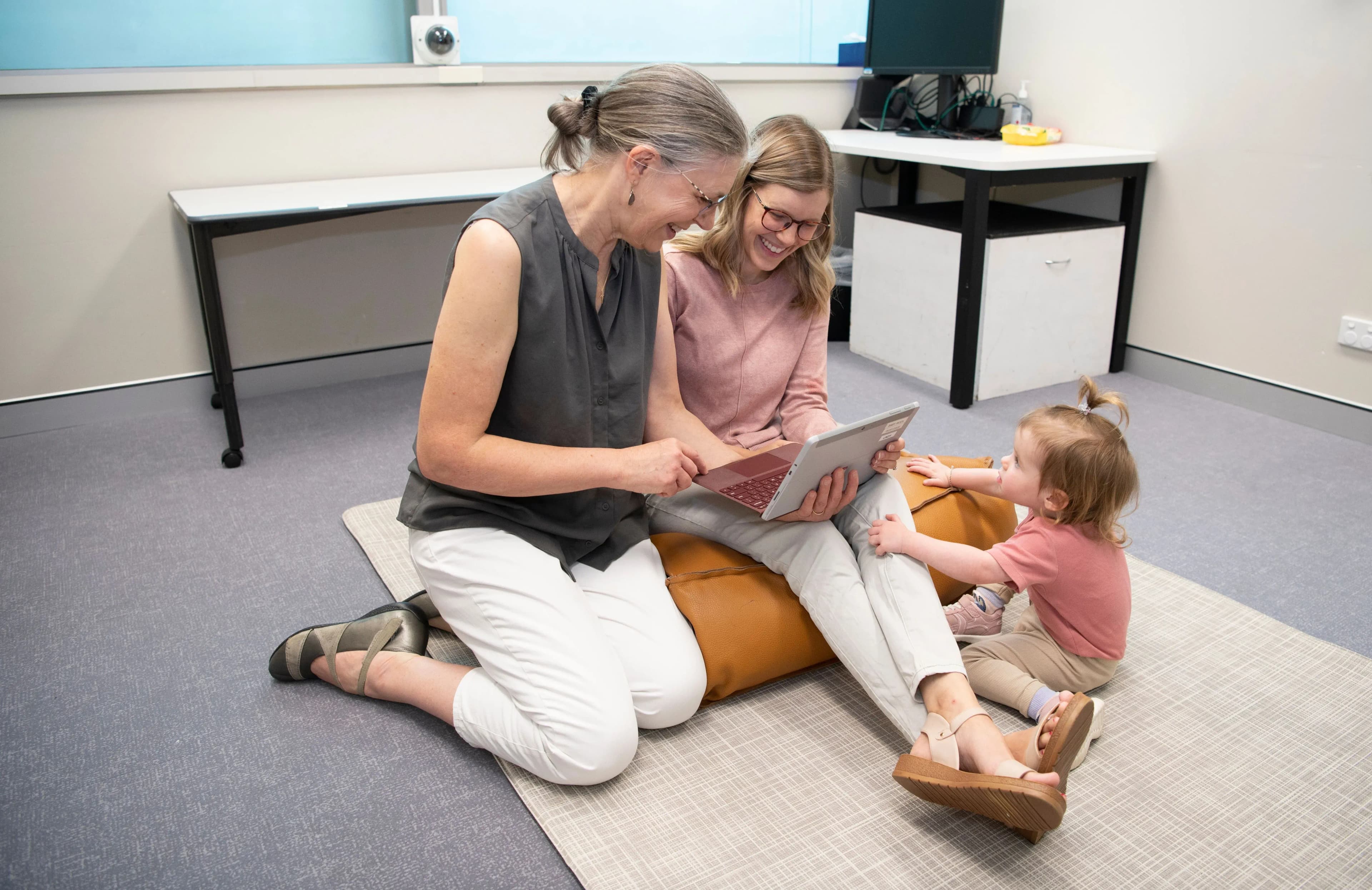 Two women sitting on the floor, discussing something they're reading on their laptop. Next to them is a child.